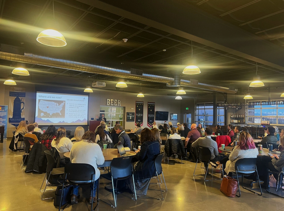 A large group of people sitting at tables in a spacious room with a presentation on a projector screen at the front. The screen displays a US map and text. Various banners are hung on the walls, and a bar is visible in the background with the word "BEER" above it.