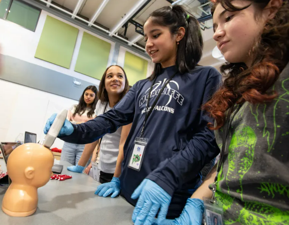 A group of girls is gathered around a table, one of them holding an ultrasound probe to a mannequin head. They are wearing gloves and appear engaged in the activity. The background features a classroom setting with pastel-colored panels on the walls.