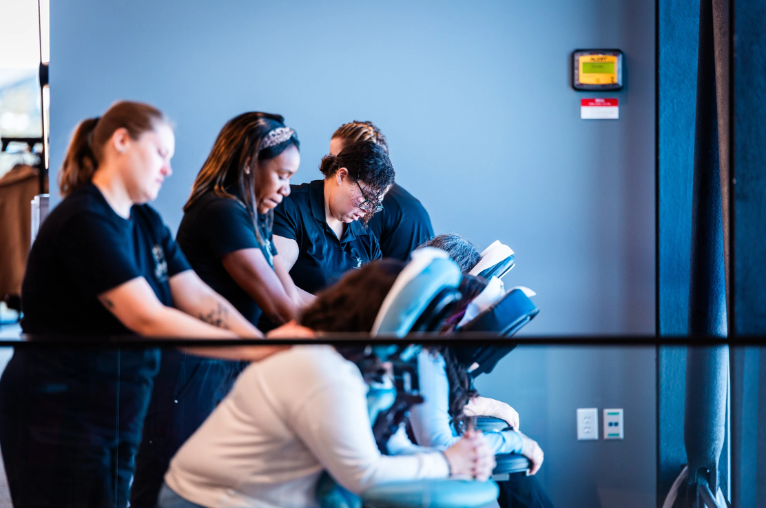 A group of people are receiving chair massages in a well-lit room. Five massage therapists work on individual clients who are sitting in specialized massage chairs. Everyone appears relaxed and focused on the massage session.