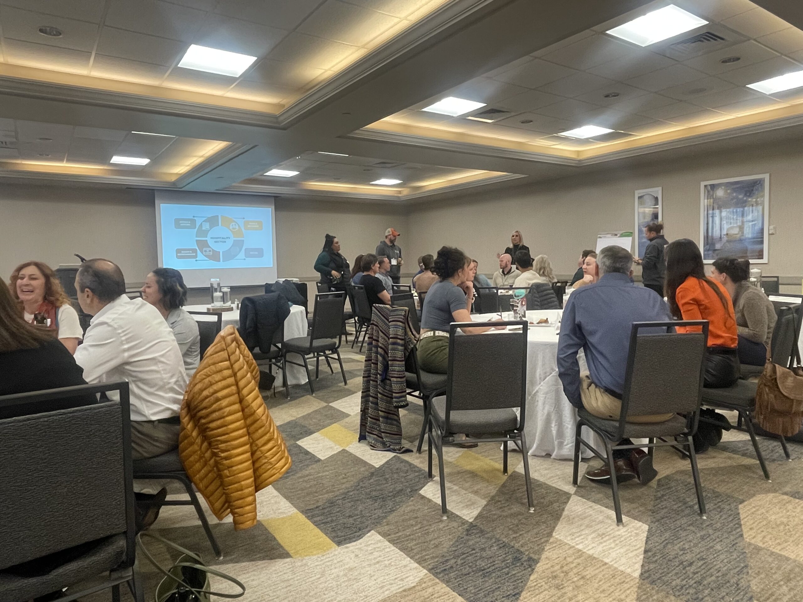 People are sitting at round tables in a conference room with a presentation on a screen. The room is well-lit, and attendees appear engaged in discussion. A yellow jacket hangs on a chair.