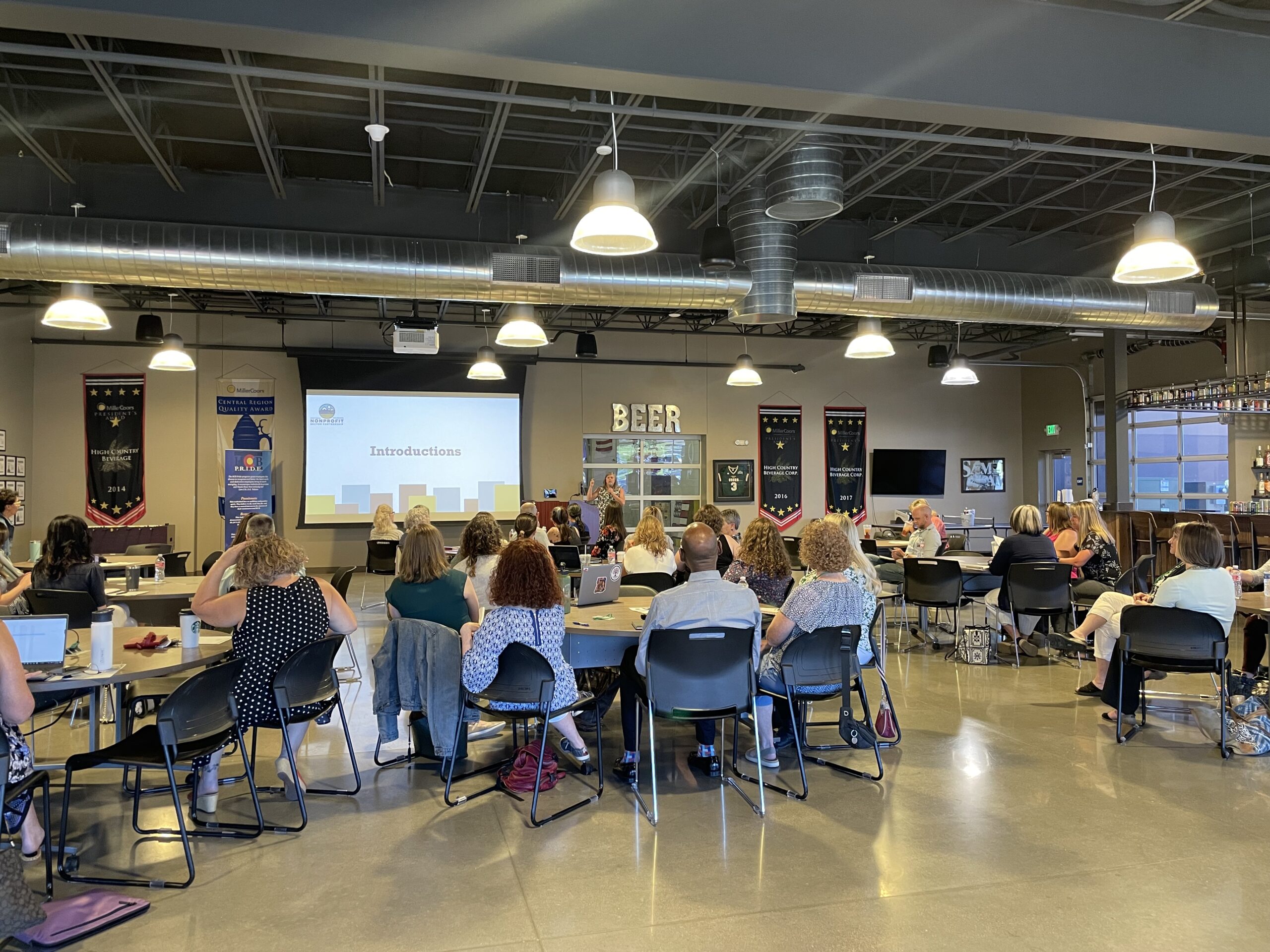 A group of people sitting at tables in a modern conference room, facing a presentation screen displaying the word "Introductions." The room has industrial lighting and a "BEER" sign in the background.