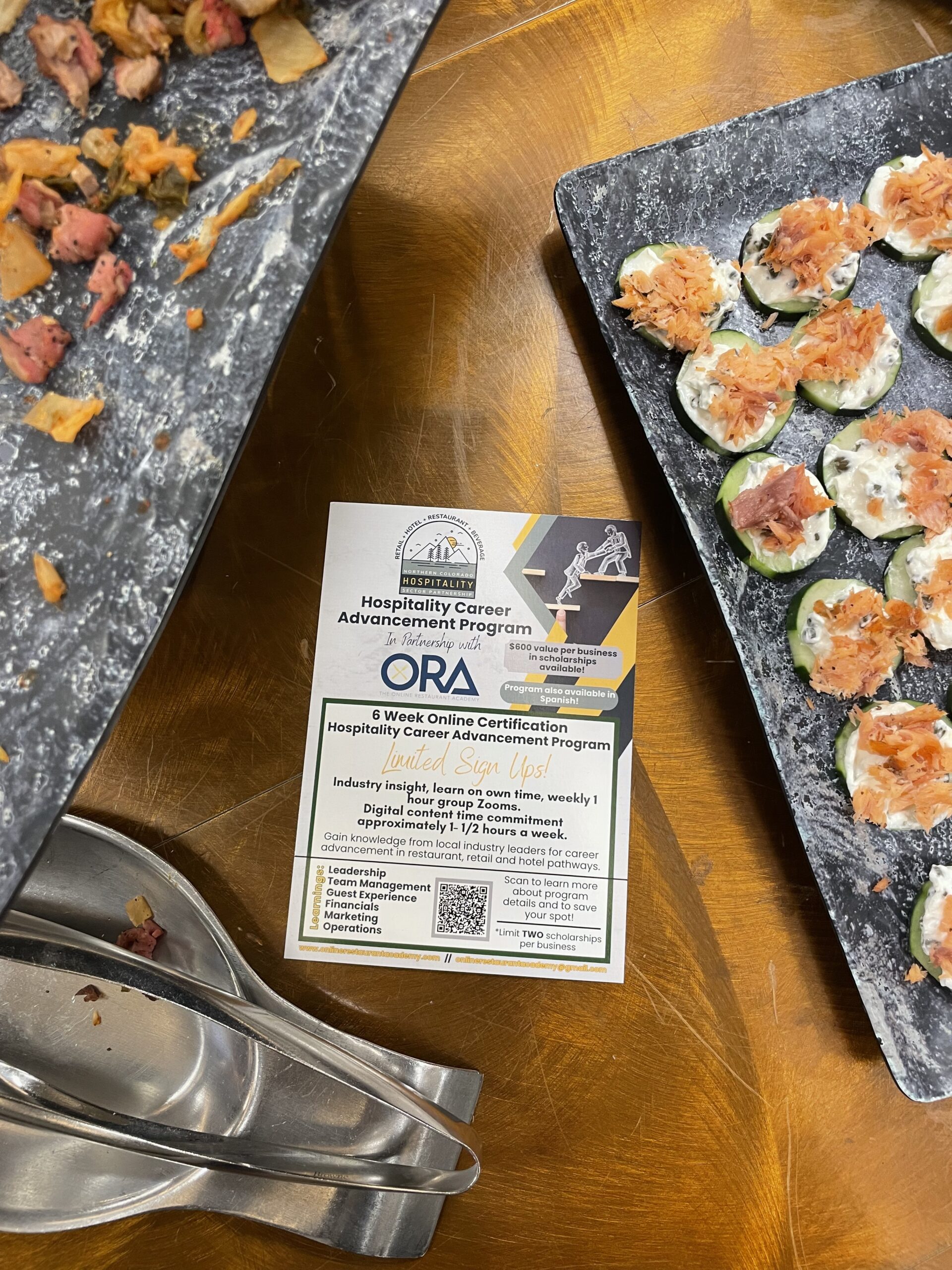 A flyer for the Hospitality Career Advancement Program is placed on a table surrounded by trays of appetizers. The flyer mentions a 6-week online certification with an option to sign up.