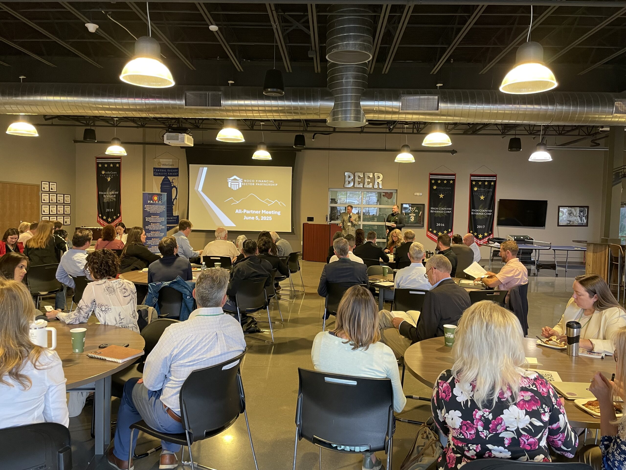 A group of people sits at round tables in a modern conference room, facing a speaker at a podium. A presentation is projected on a screen, and banners and a "BEER" sign are visible on the walls.