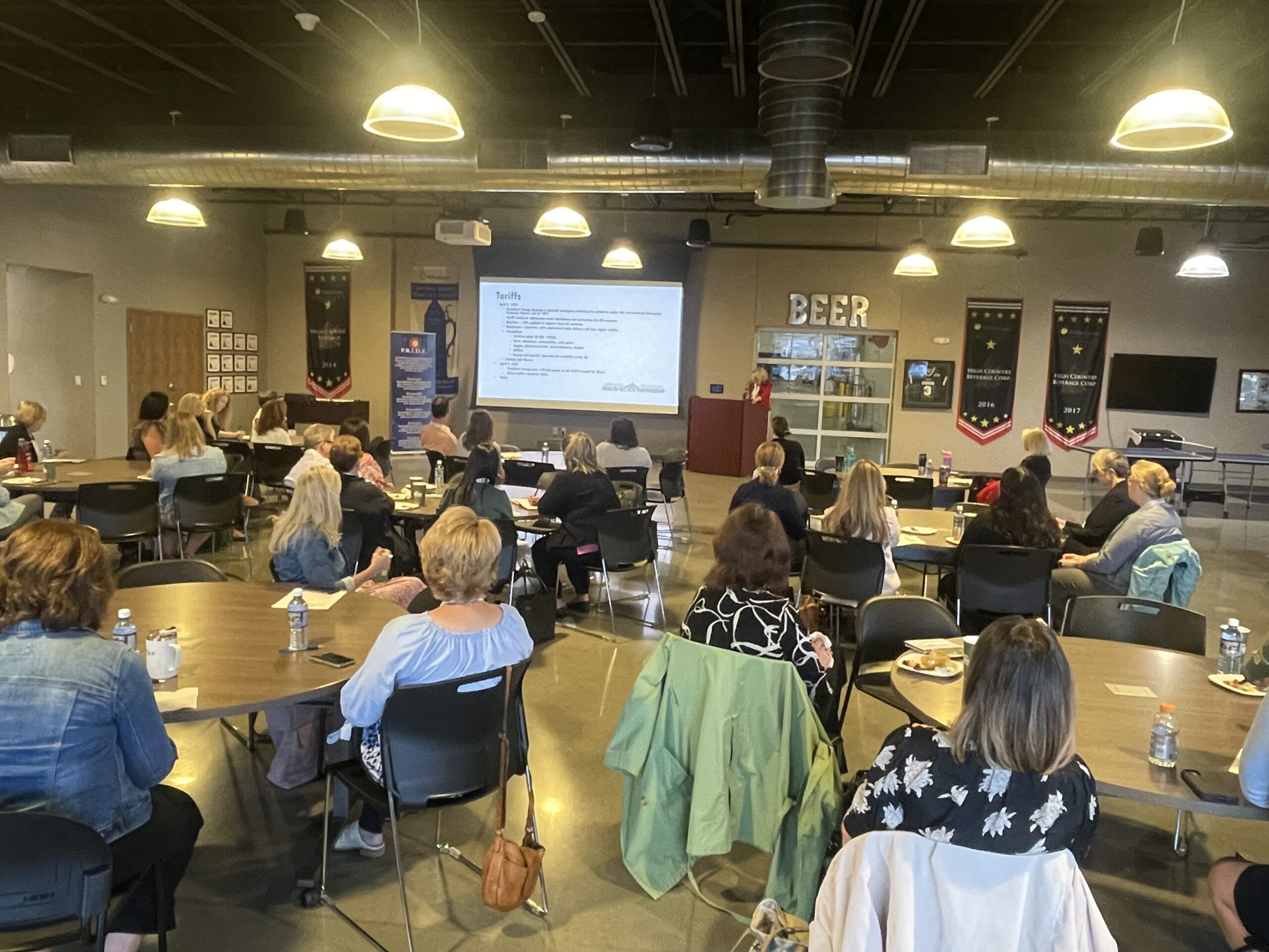 A group of people sits at round tables in a large room, watching a presentation projected on a screen at the front. The room has high ceilings, banners, and exposed ductwork.
