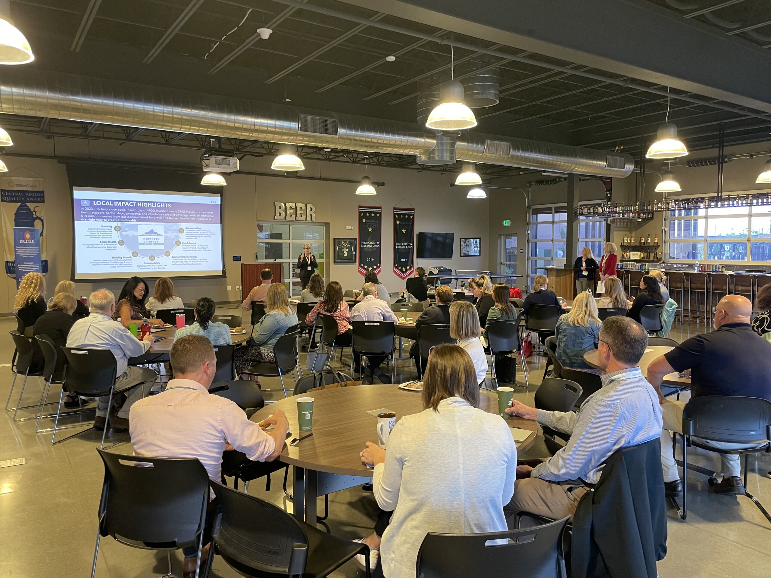 A group of people sit at round tables in a large room, attentively listening to a speaker presenting slides at the front. The room has high ceilings, hanging lights, and “BEER” spelled out on the back wall.