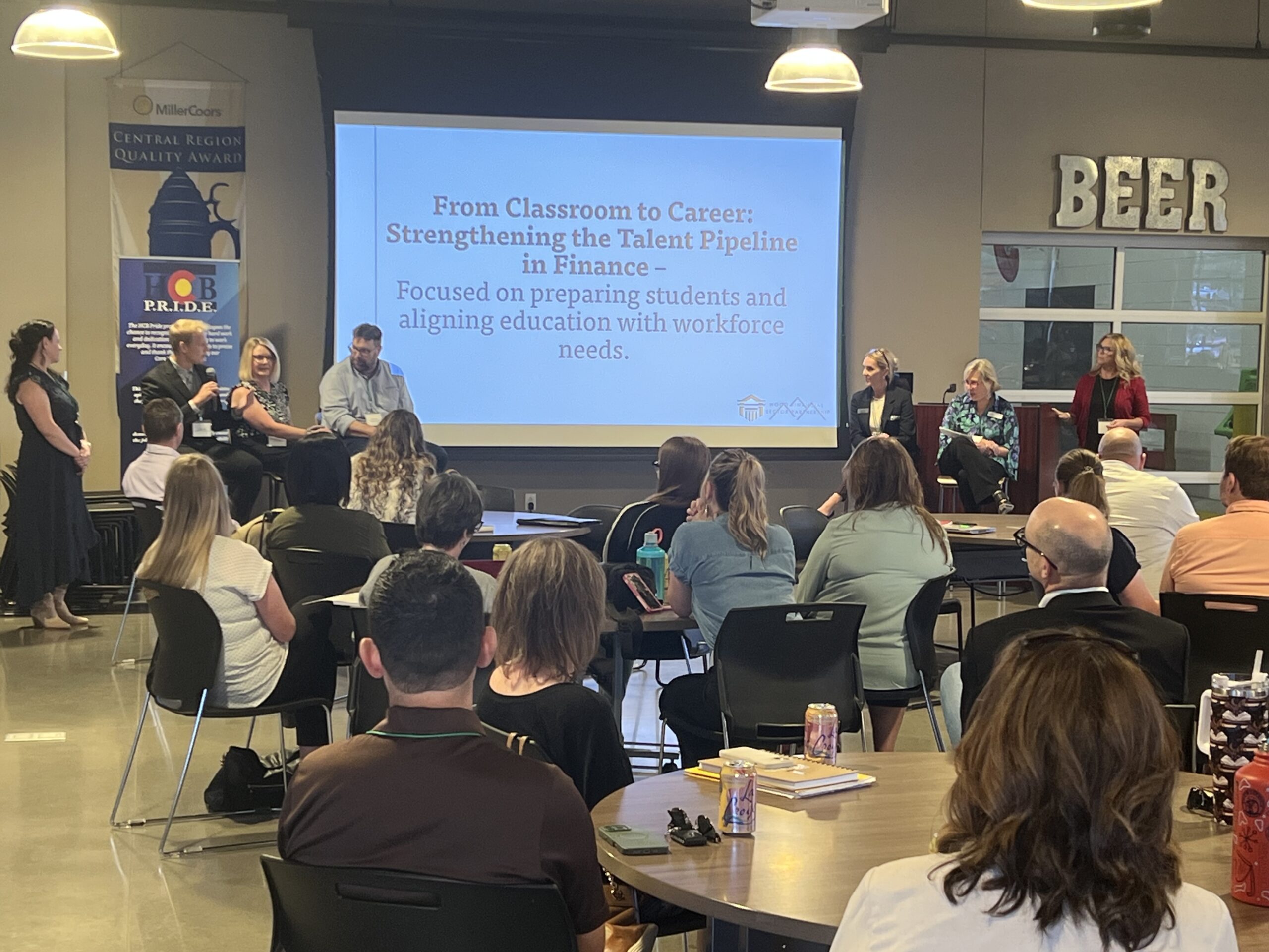 A panel of speakers sits on a stage in front of a projected presentation that reads, "From Classroom to Career: Strengthening the Talent Pipeline in Finance." Audience members are seated and listening attentively.