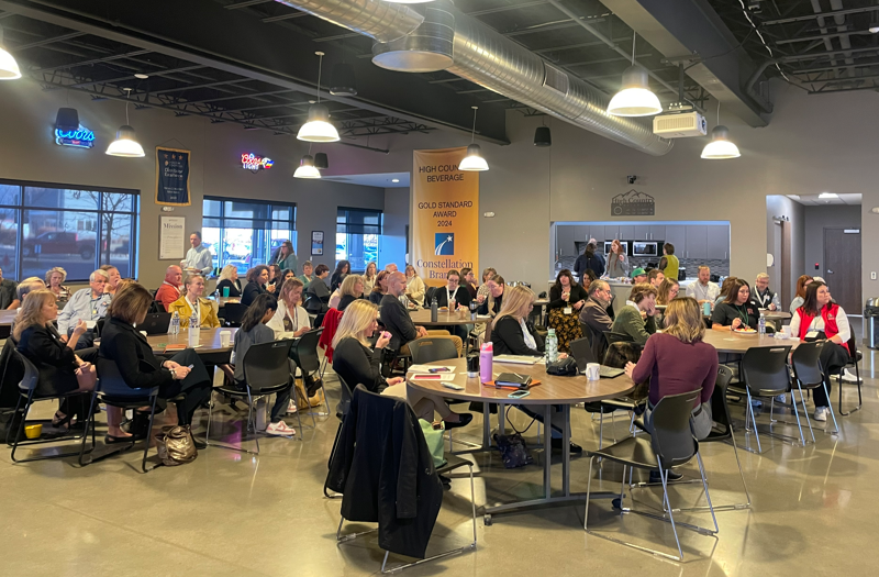 A group of people sit at round tables in a modern, open room, listening to a speaker at the front. The room has industrial-style ceilings, large windows, and bright lighting.