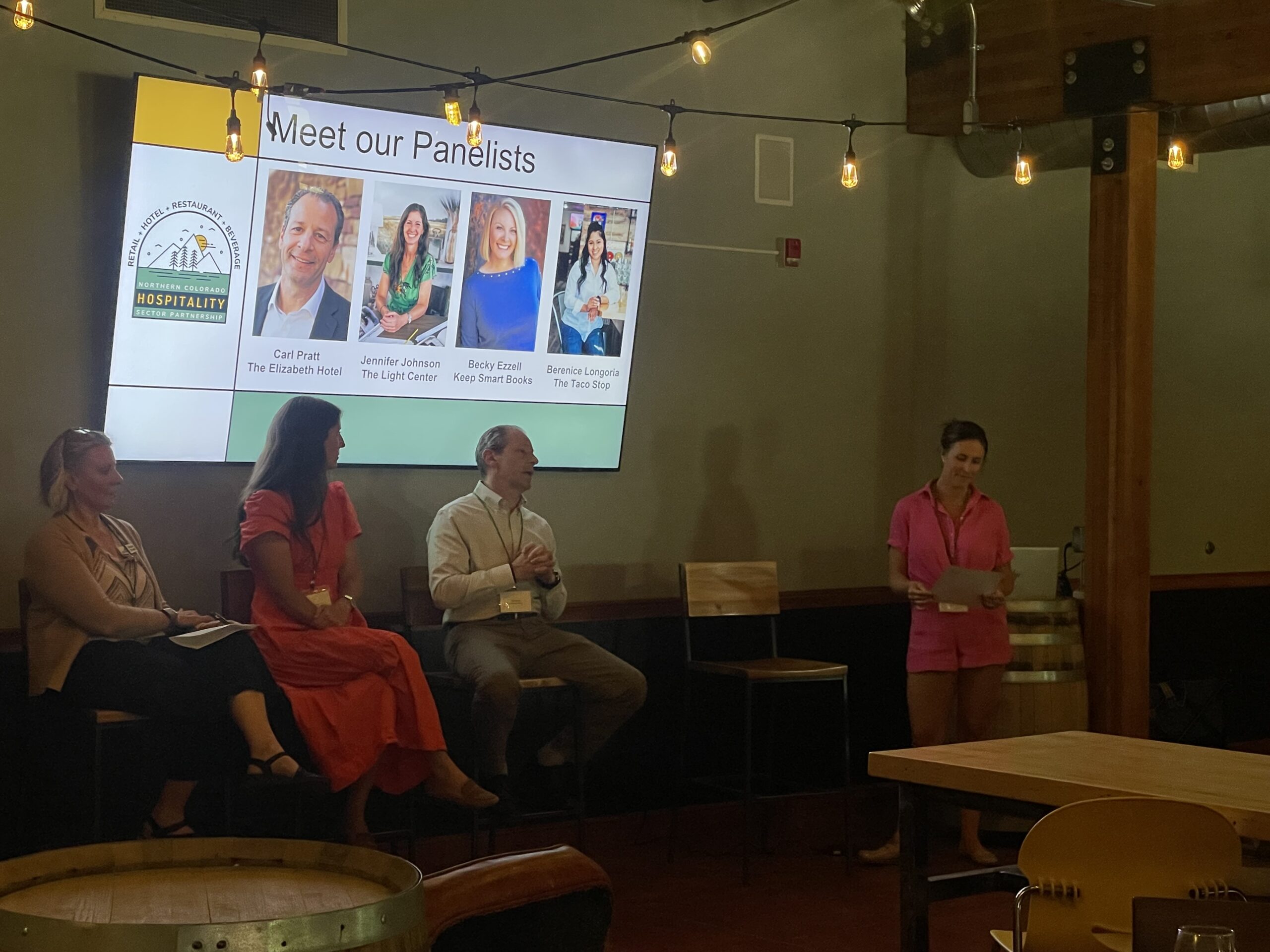 A woman in pink stands and speaks near four panelists seated under string lights, with a screen behind them displaying "Meet our Panelists" and their names and photos.