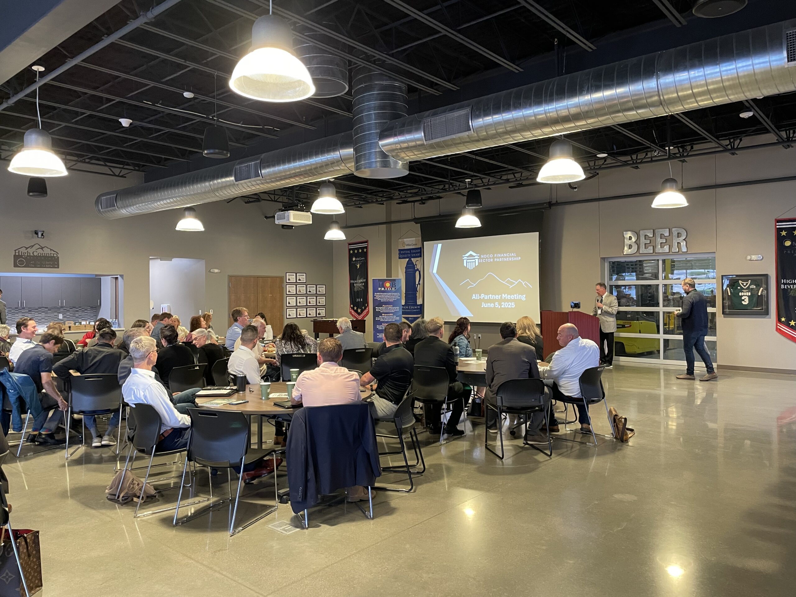 A group of people sit at round tables in a modern, industrial-style room, listening to a speaker at a podium with a presentation projected on a screen. Signs and sports memorabilia decorate the walls.