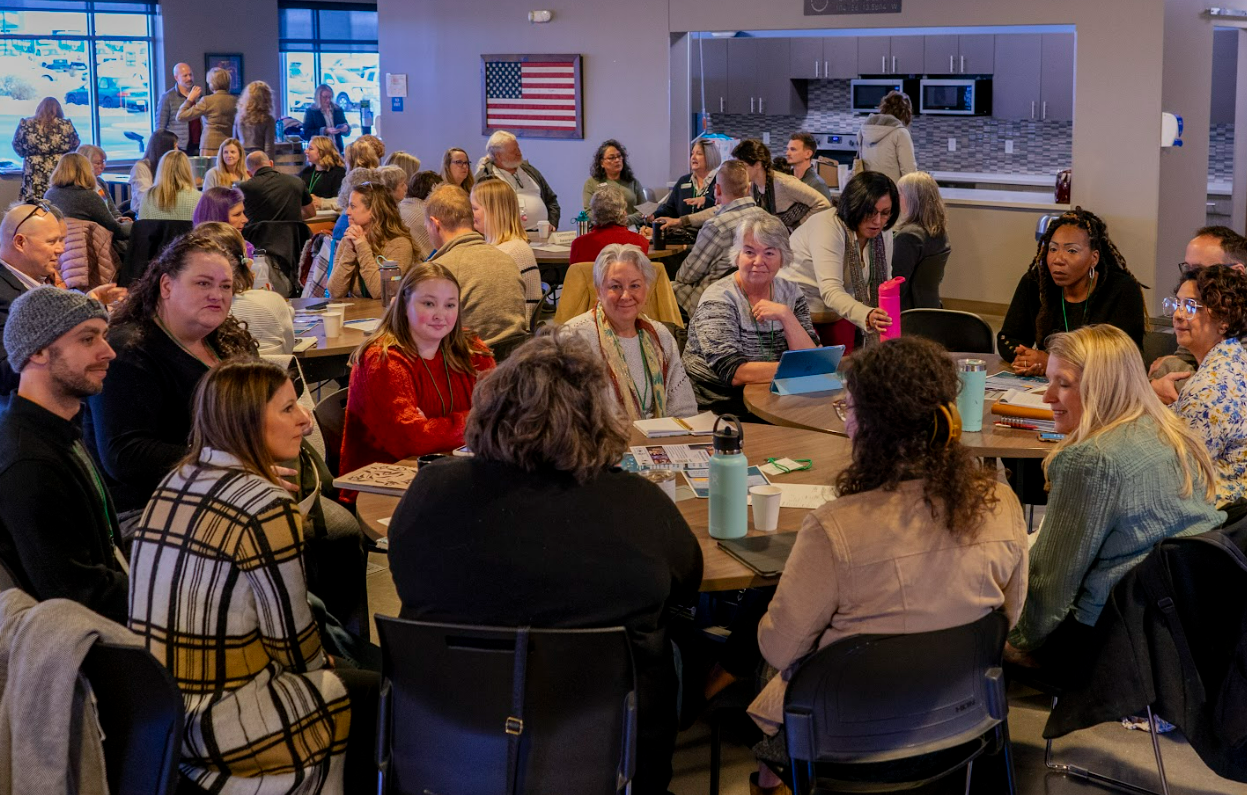 A large group of people sit around tables in a busy, well-lit room, engaged in conversation. The atmosphere appears friendly and collaborative, with diverse individuals and an American flag on the wall in the background.