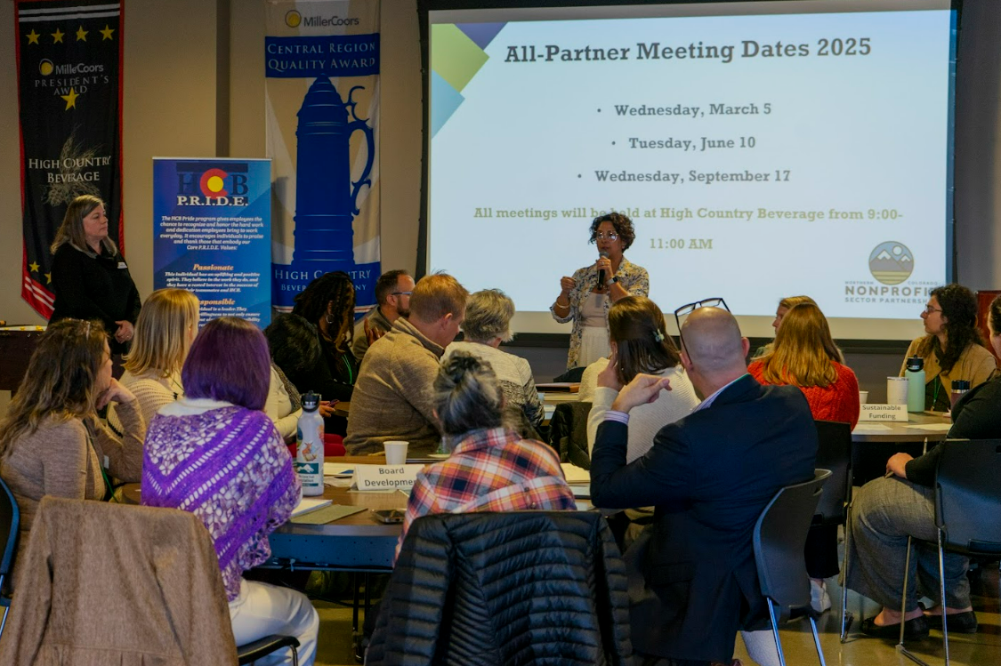 A speaker addresses a group of attendees seated at tables in front of a projection screen displaying 2025 meeting dates.