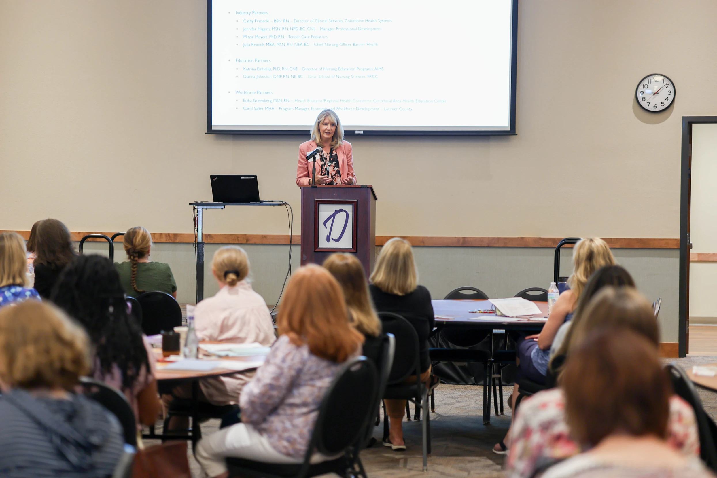 A woman in a pink blazer speaks from a podium to an audience in a conference room, with a projector screen listing “Industry Partners,” “Education Partners,” and “Workforce Partners” behind her.